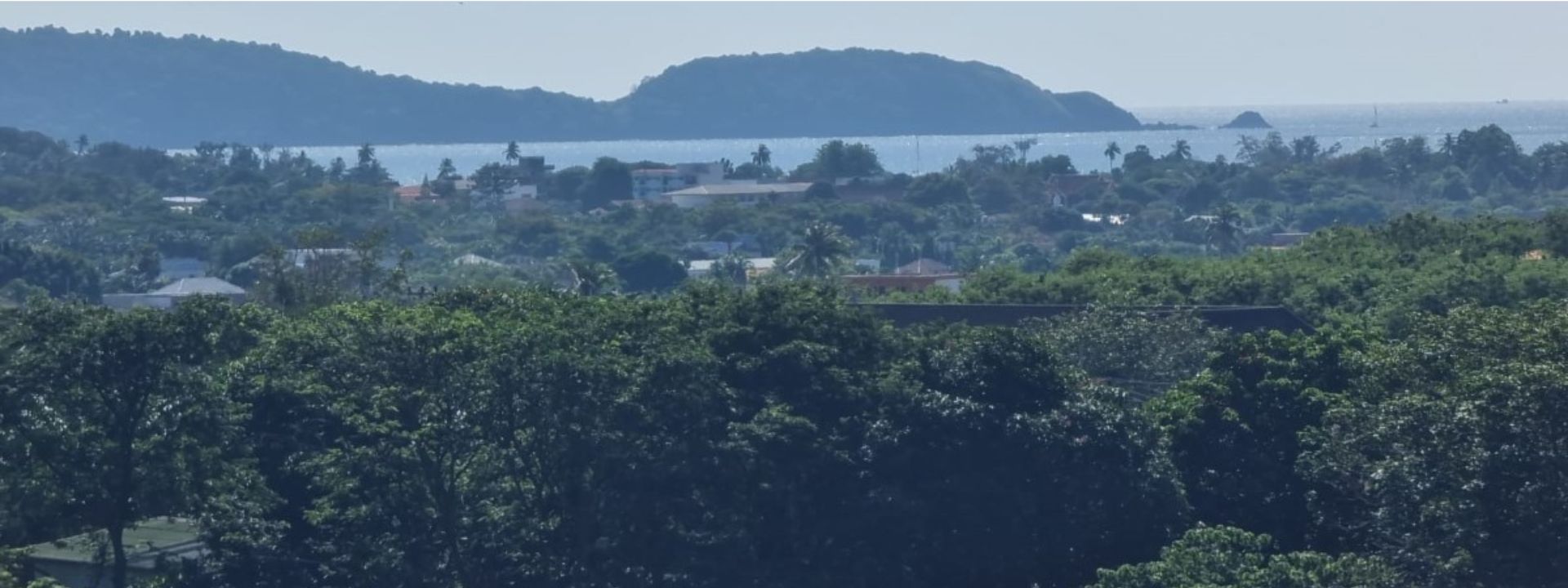 Coastal hillside view in Phuket with dense greenery, low-rise homes and an island in the distance.