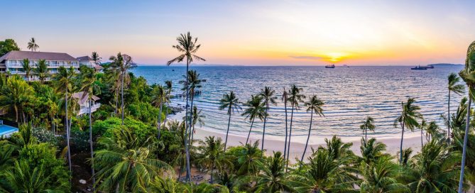 Sunset view of Panwa Beach in Phuket with palm trees, calm sea and beachfront resorts.