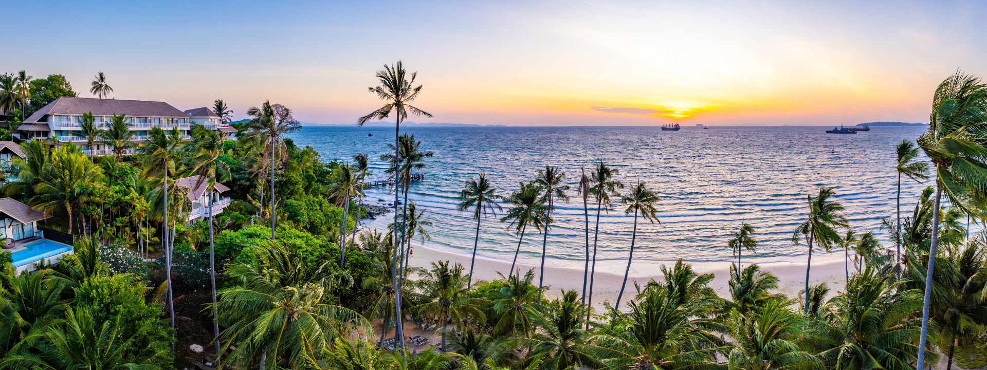Sunset view of Panwa Beach in Phuket with palm trees, calm sea and beachfront resorts.