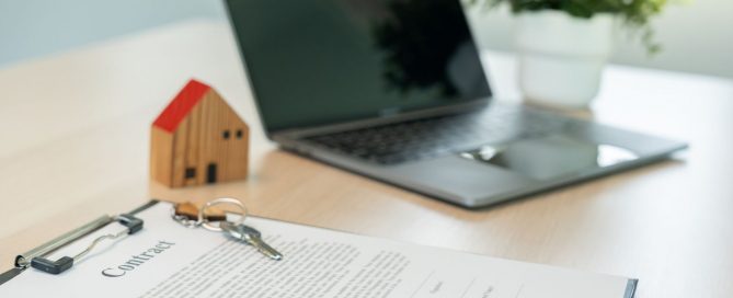 Real estate contract with house keys, miniature house model, and laptop on a desk.