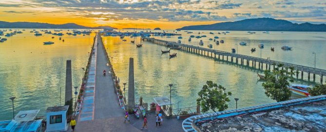 Sunset view of Chalong Pier in Phuket with boats, calm water and surrounding residential coastline.