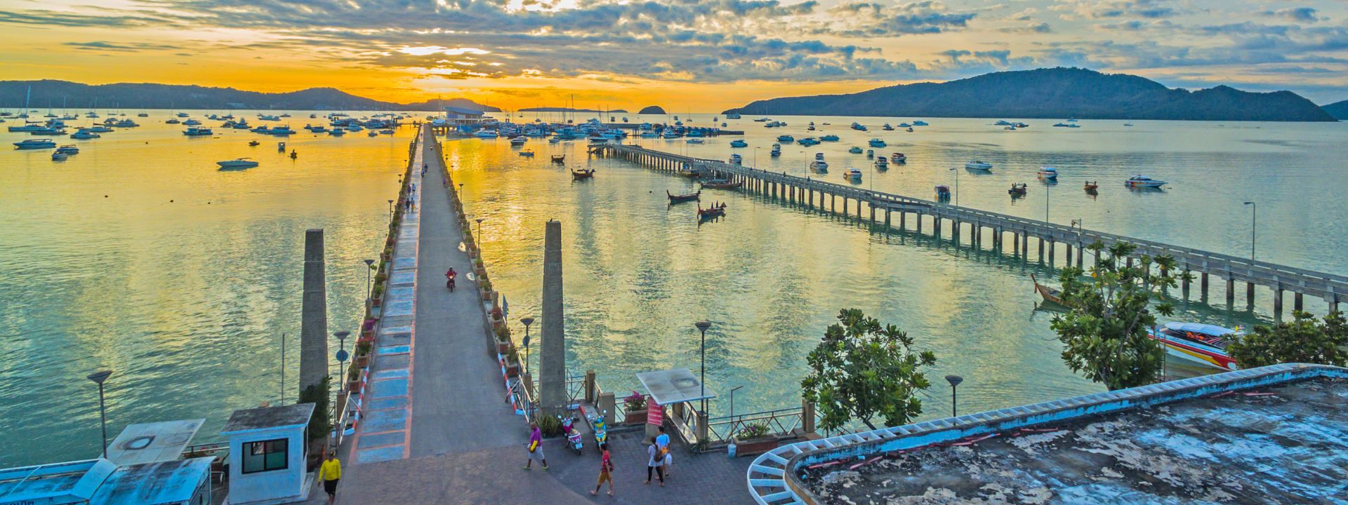 Sunset view of Chalong Pier in Phuket with boats, calm water and surrounding residential coastline.
