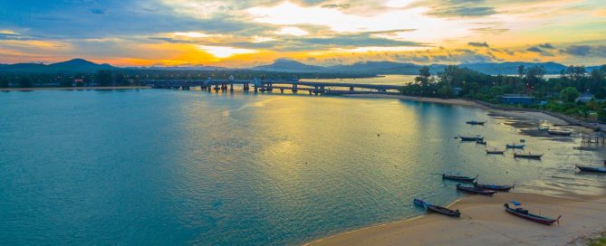 Sunset view of Sarasin Bridge in Phuket with calm water, longtail boats and coastal scenery.