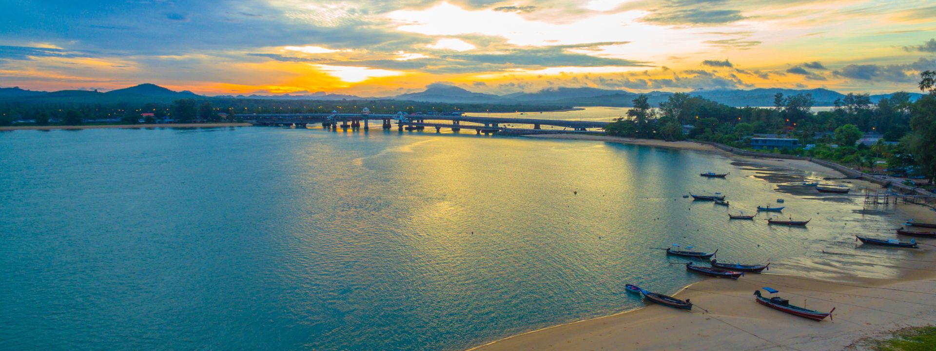 Sunset view of Sarasin Bridge in Phuket with calm water, longtail boats and coastal scenery.