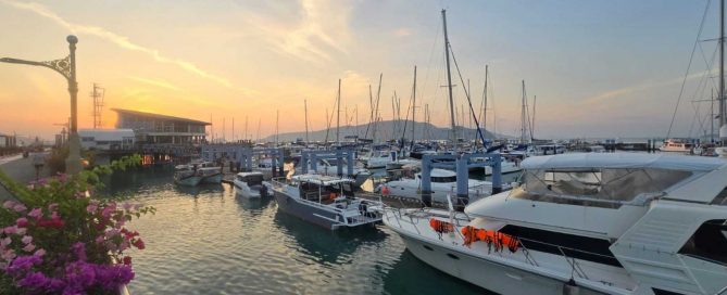 Sunrise over Chalong Bay in Phuket with luxury yachts and sailboats moored at the marina, calm water reflecting the soft morning sky.