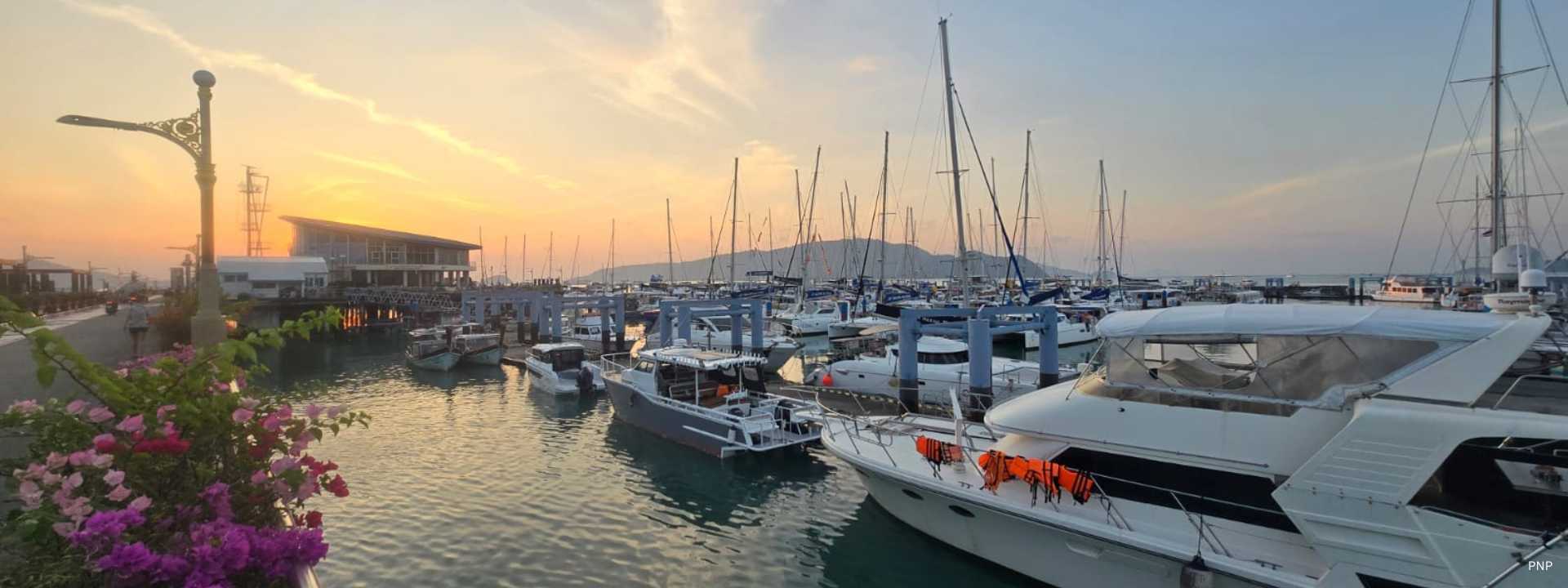Sunrise over Chalong Bay in Phuket with luxury yachts and sailboats moored at the marina, calm water reflecting the soft morning sky.