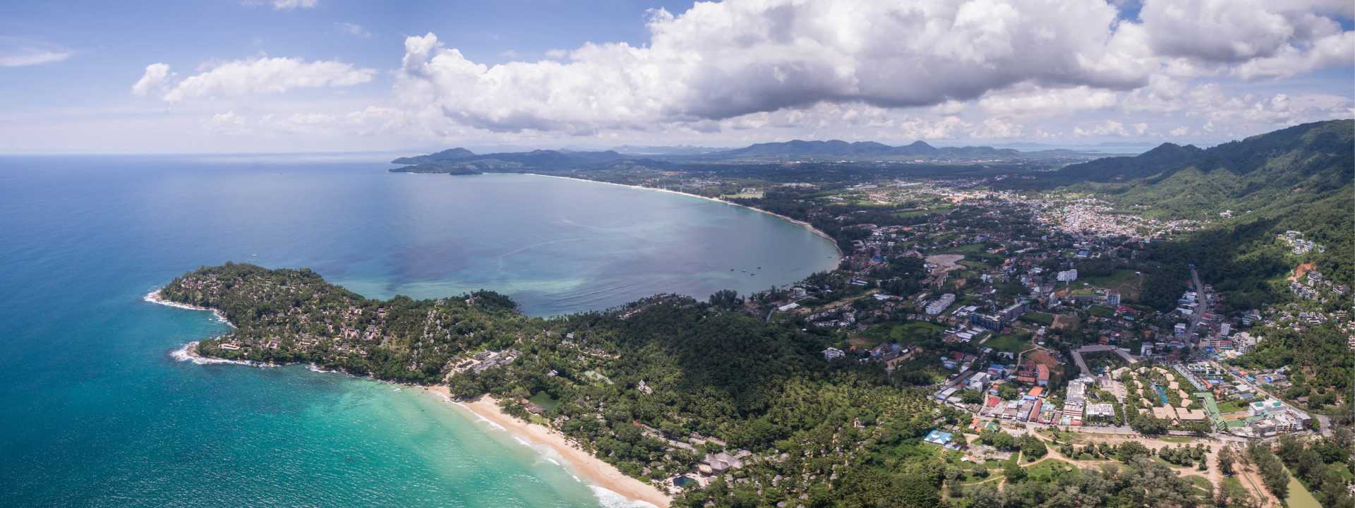 Aerial view of Bang Tao coastline in Phuket showing the curved beach, turquoise sea, headland, and surrounding town and hills.
