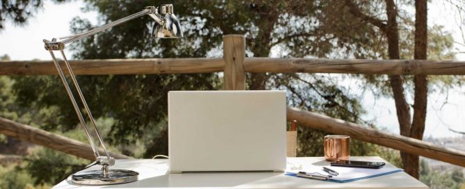 Outdoor workspace with laptop, desk lamp, notebook and coffee overlooking nature
