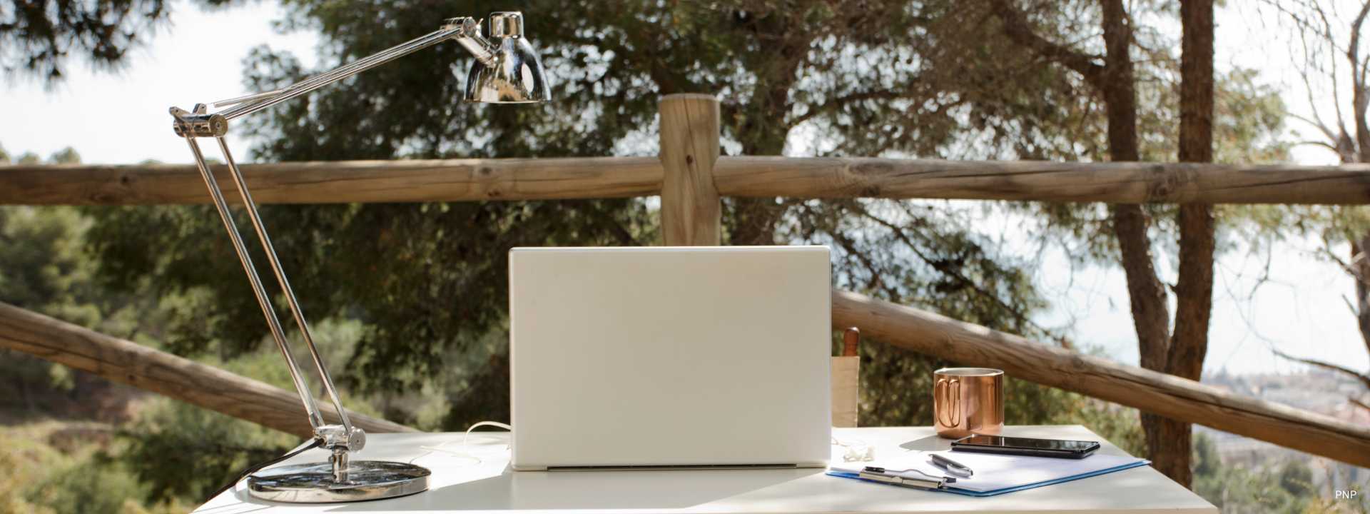 Outdoor workspace with laptop, desk lamp, notebook and coffee overlooking nature