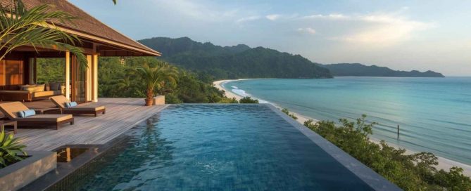 Luxury hillside villa in Phuket with a private infinity pool overlooking a tropical bay, white sand beach, and turquoise ocean at sunset.