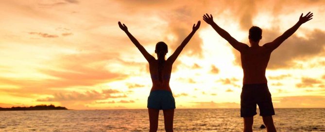 Couple standing on a Phuket beach at sunset with arms raised, enjoying coastal lifestyle