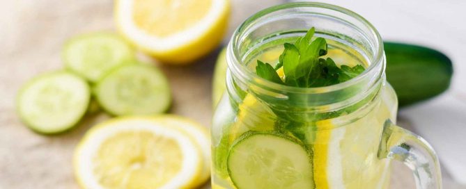 Glass jar of cucumber and lemon infused water with fresh herbs on a light surface