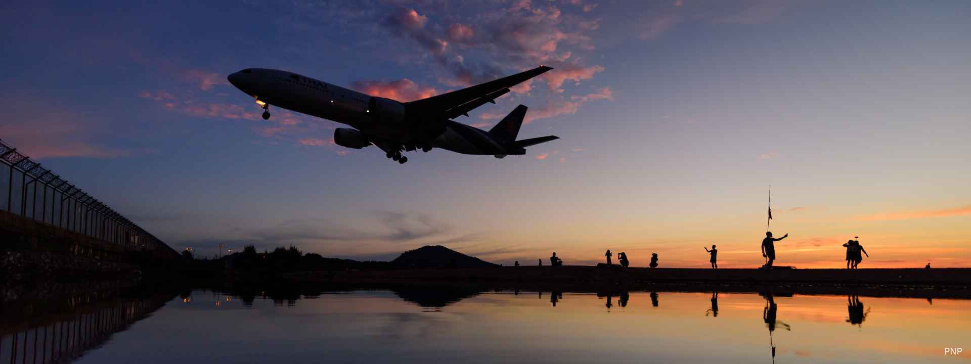 Airplane landing over Mai Khao Beach in Phuket at sunset with silhouettes of people watching and colourful evening sky