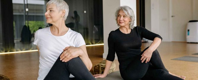 Two older women practising gentle stretching exercises on yoga mats inside a fitness studio