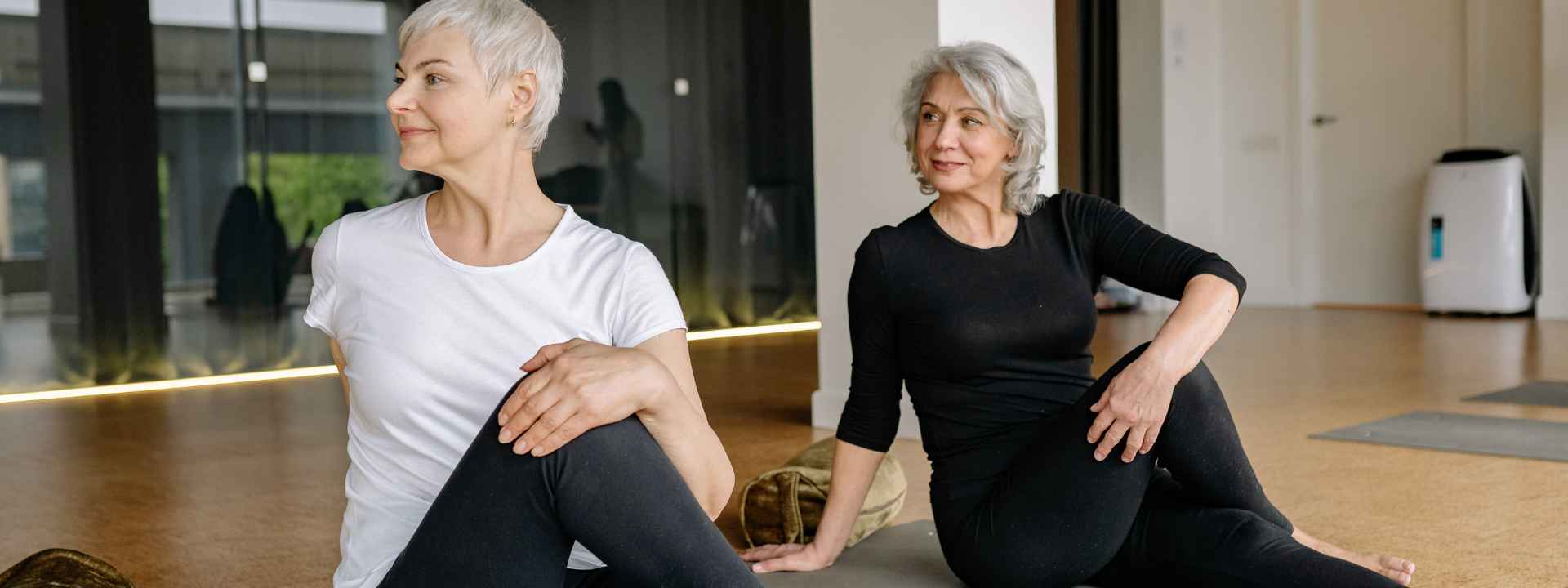 Two older women practising gentle stretching exercises on yoga mats inside a fitness studio