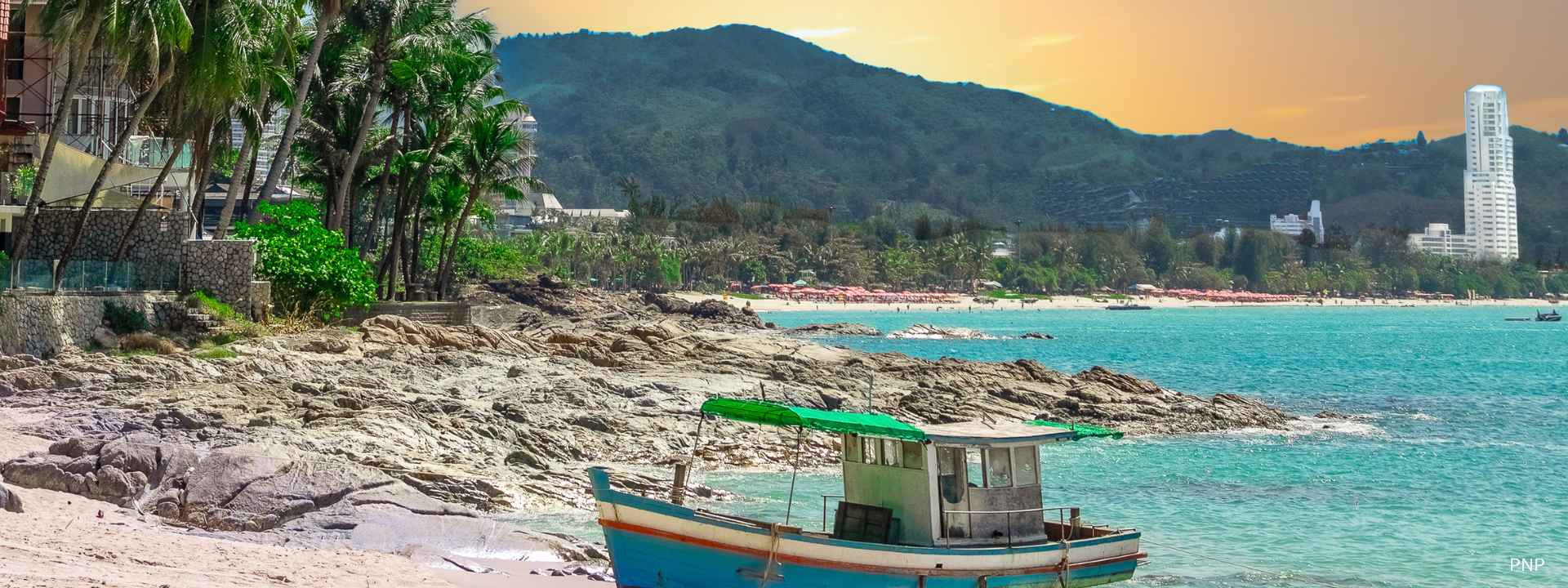 Traditional longtail boat on the rocky shoreline of Patong Beach, Phuket, with turquoise sea, palm trees, and hillside skyline at sunset