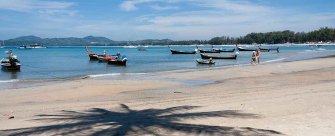 Longtail boats anchored along a sandy beach in Phuket with calm blue sea, distant hills, and palm tree shadow in the foreground