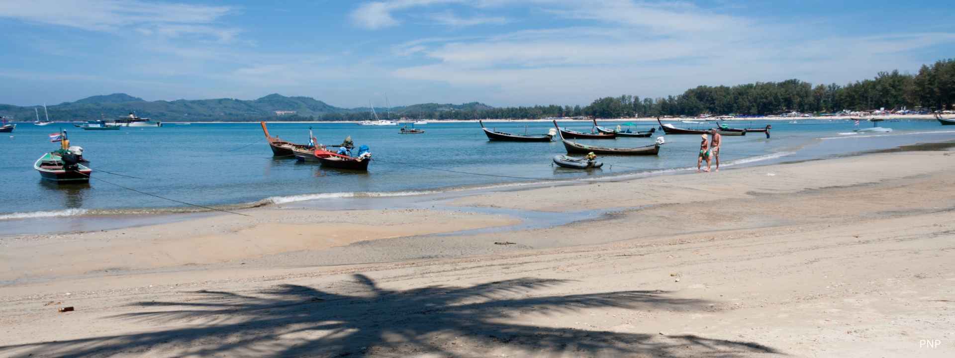 Longtail boats anchored along a sandy beach in Phuket with calm blue sea, distant hills, and palm tree shadow in the foreground