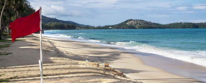 Red warning flag on a sandy Phuket beach with turquoise sea, gentle waves, and palm-lined shoreline