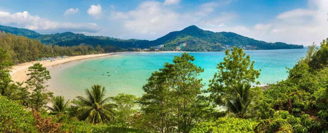 Panoramic view of a tropical beach in Phuket with turquoise water, sandy shoreline, palm trees, and green hills in the background