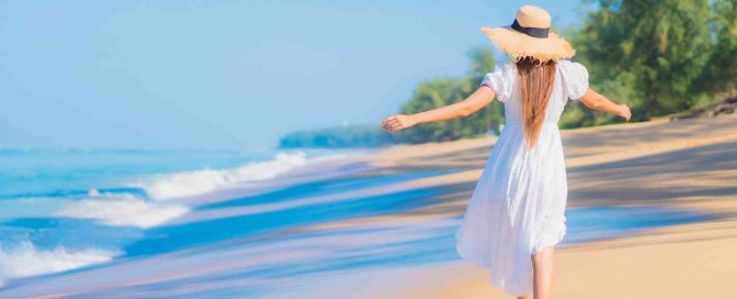 Woman in a white dress walking along a tropical beach in Phuket with blue sea, gentle waves, and palm trees in the background