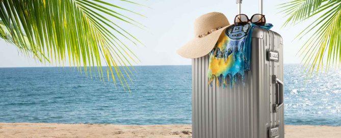 Suitcase with sun hat and beach towel on a sandy Phuket beach with palm leaves and blue ocean in the background
