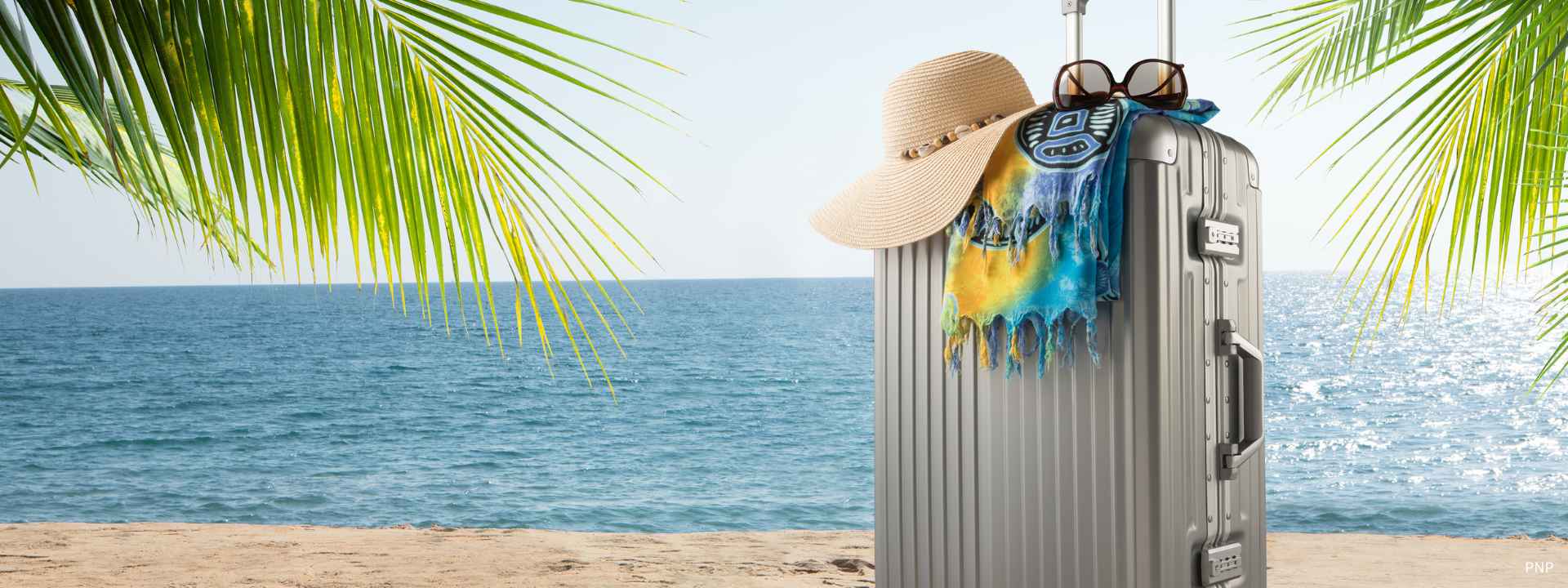 Suitcase with sun hat and beach towel on a sandy Phuket beach with palm leaves and blue ocean in the background