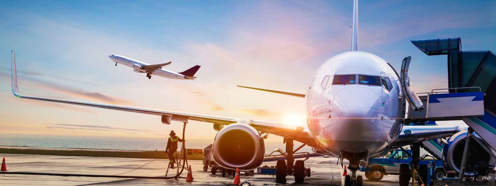Commercial airplane being refueled on an airport runway at sunrise with another plane taking off in the background