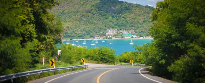 Coastal road in Phuket overlooking a bay with boats and hillside property development in the background