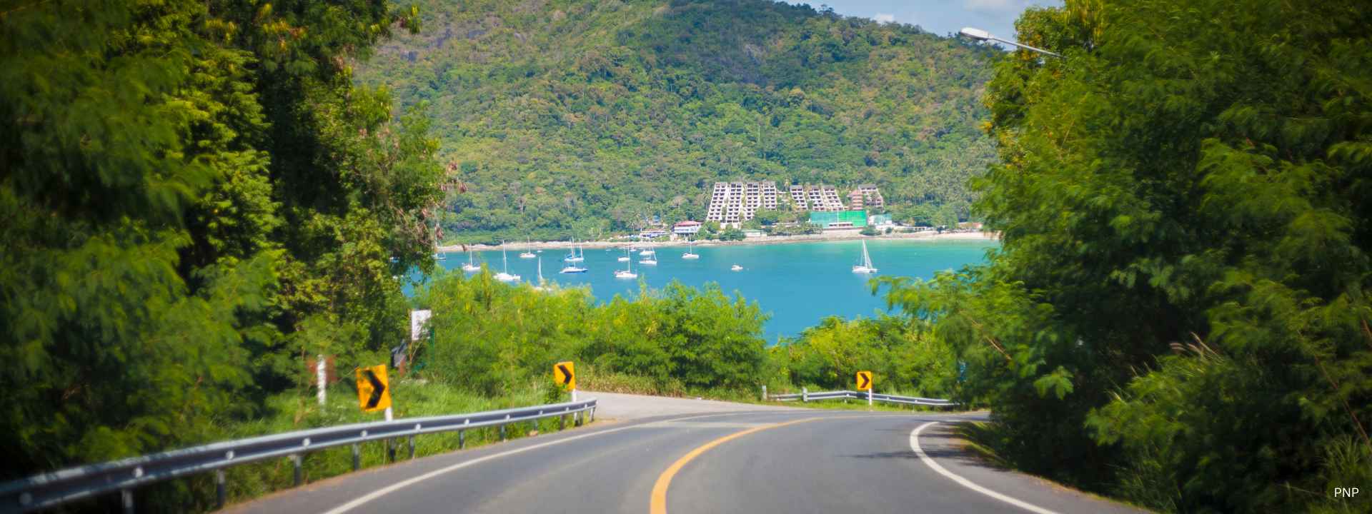 Coastal road in Phuket overlooking a bay with boats and hillside property development in the background
