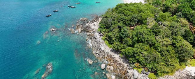 Aerial view of Phuket's tropical coastline with lush rainforest, granite rocks, turquoise Andaman Sea waters, and traditional longtail boats.