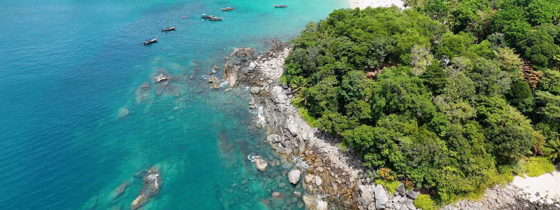 Aerial view of Phuket's tropical coastline with lush rainforest, granite rocks, turquoise Andaman Sea waters, and traditional longtail boats.