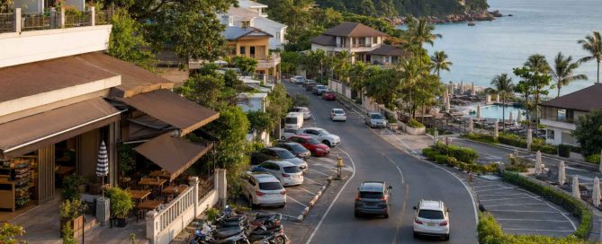 Vehicles and parked motorbikes line an active Phuket coastal road beside quiet resort pool terraces overlooking the Andaman Sea.
