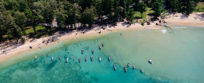 Aerial view of Layan Beach in Phuket with traditional longtail boats anchored in clear turquoise water, highlighting the peaceful neighbourhood and natural beauty of Layan.