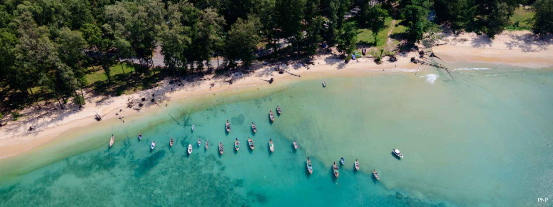 Aerial view of Layan Beach in Phuket with traditional longtail boats anchored in clear turquoise water, highlighting the peaceful neighbourhood and natural beauty of Layan.